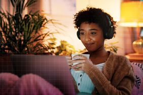 a photo of a woman sitting on the couch with a drink and her laptop and headphones at night 