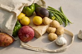 An assortment of vegetables and fruits spilling out of a reusable bag onto a flat surface, including avocados, lemons, potatoes, and more