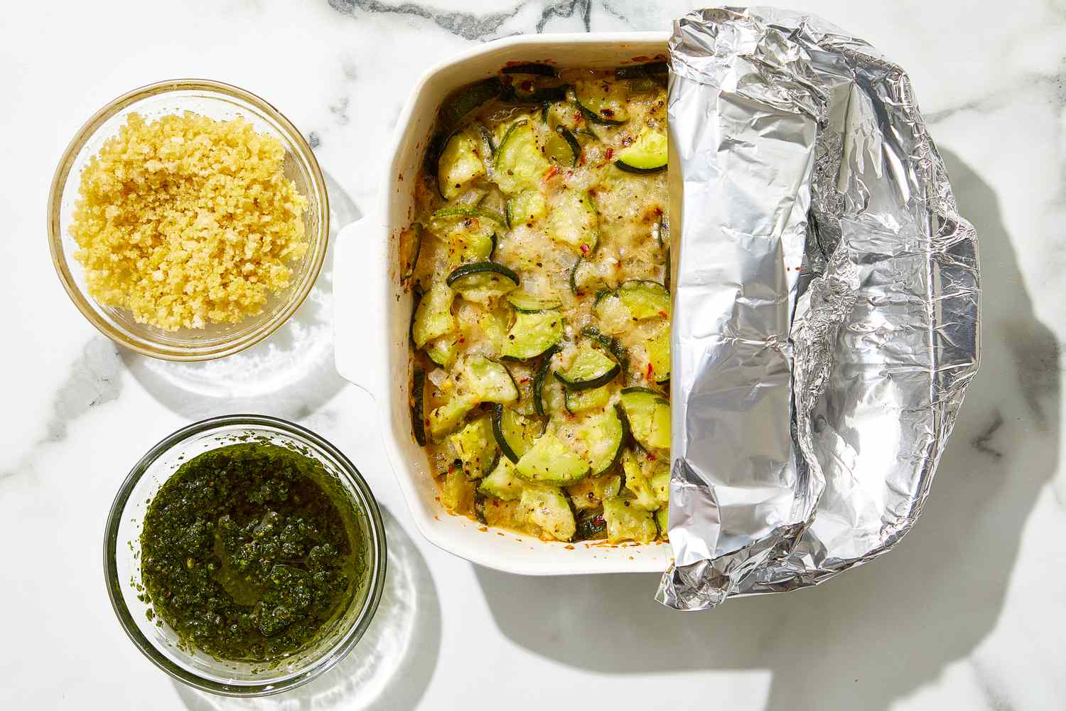 overhead view of casserole half covered with foil, a bowl of pesto, and a bowl of panko