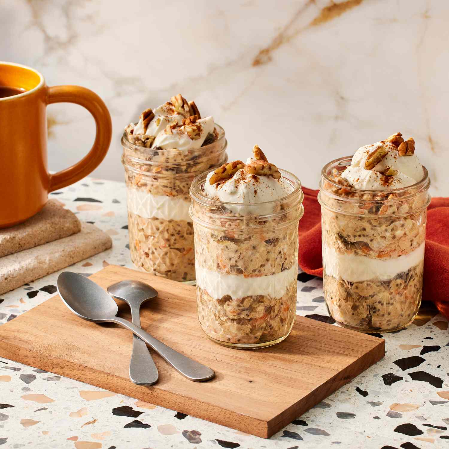 three small jars of carrot cake overnight oats, two spoons, a cloth napkin and a cup of coffee