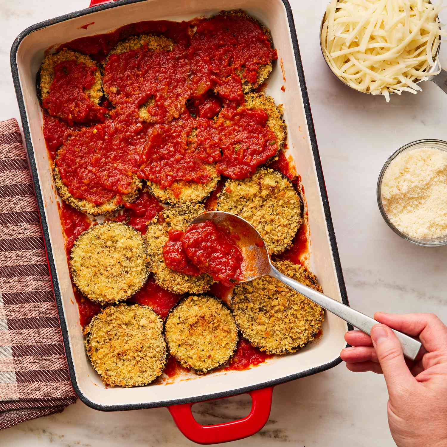 panko crusted, baked eggplant slices in a baking dish with a hand spreading tomato sauce on top, a cup of shredded mozzarella and a cup of grated Parmesan