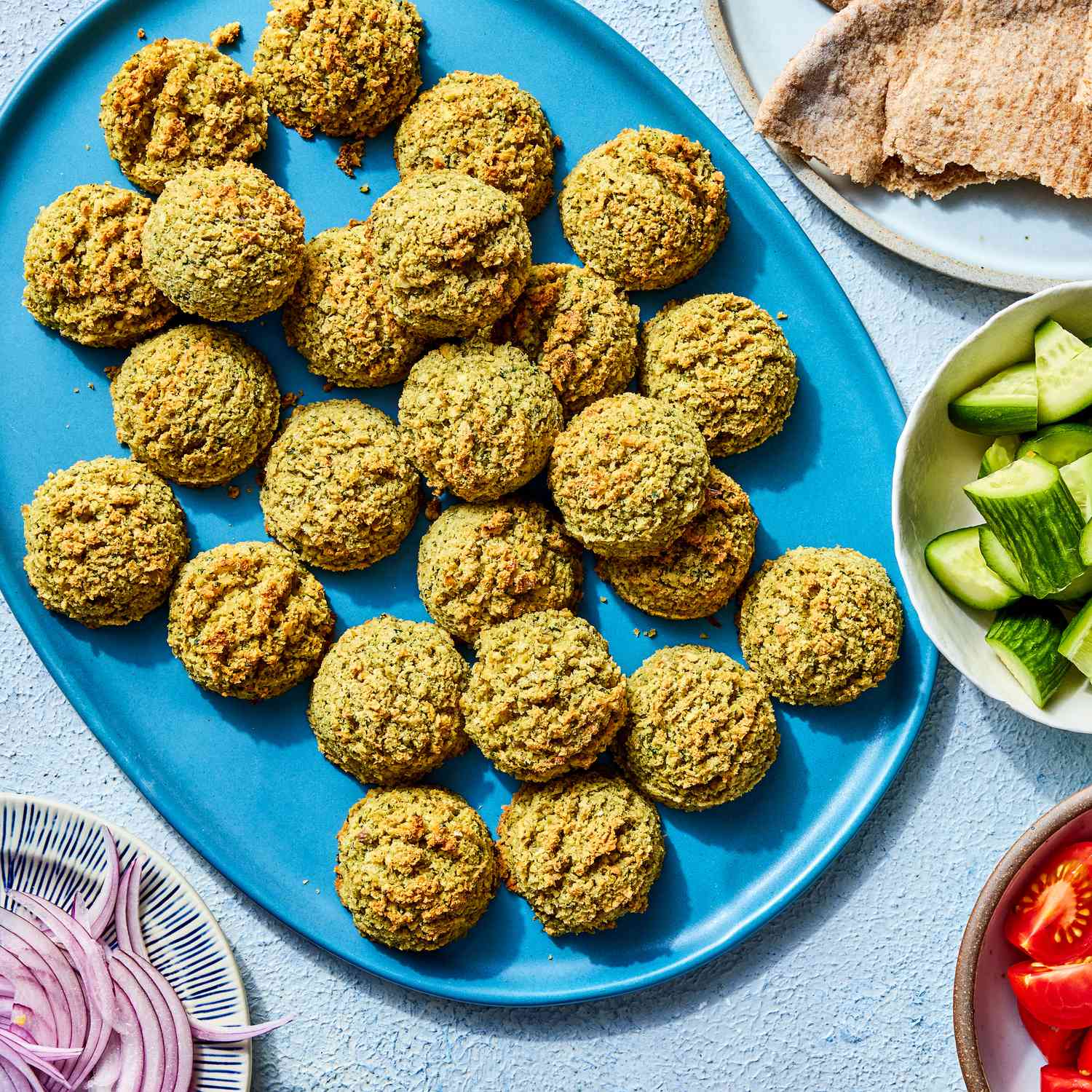 Baked falafel on a bright blue serving plate, with some pita bread, chopped cucumber, tomatoes and onions on the side