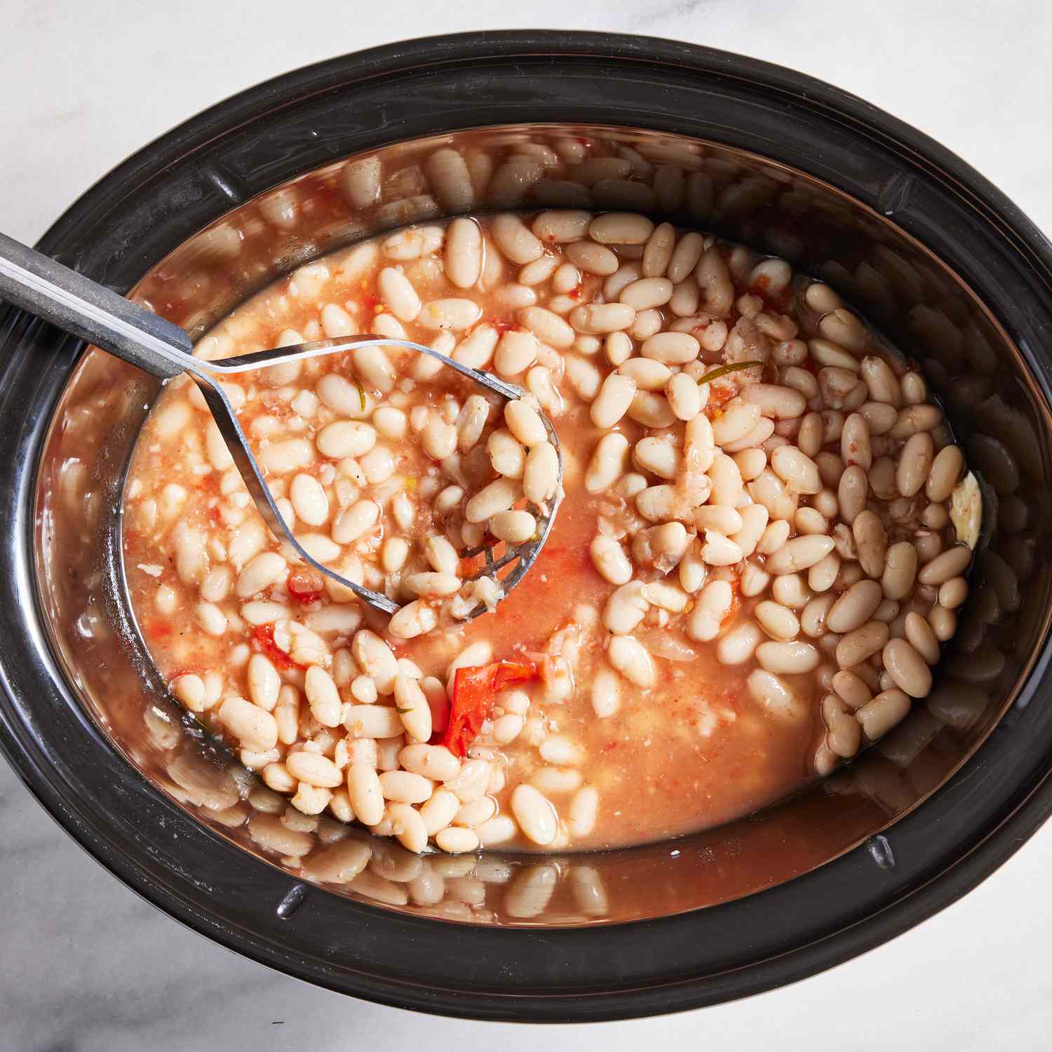 White beans in a slow-cooker getting mashed with a potato masher