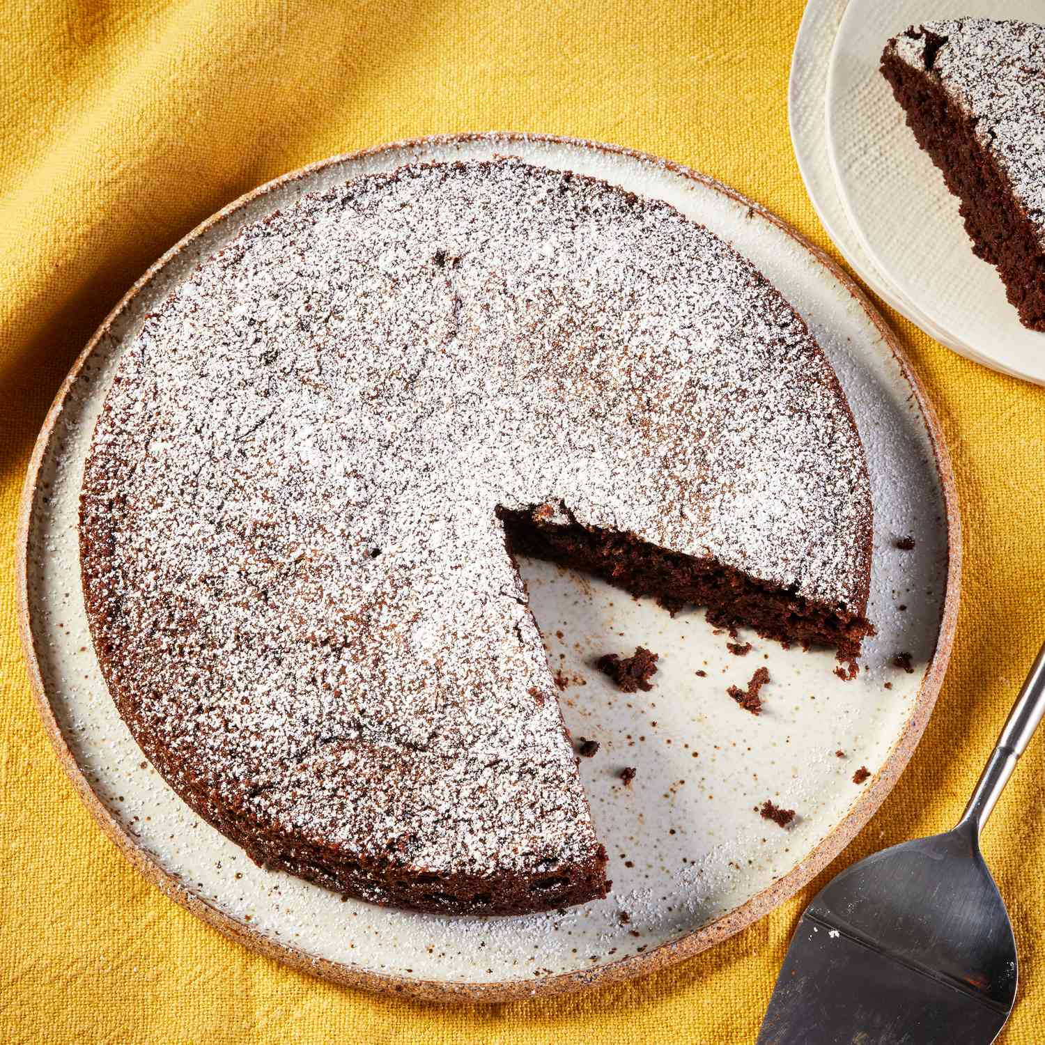 One-bowl chocolate cake, on a white plate on a yellow cloth background, with one slice of the cake cut out and put on another cake
