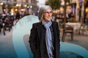 A woman with gray hair and glasses walks outdoors on a city street, hands in pockets, blurred background with lights and tables.